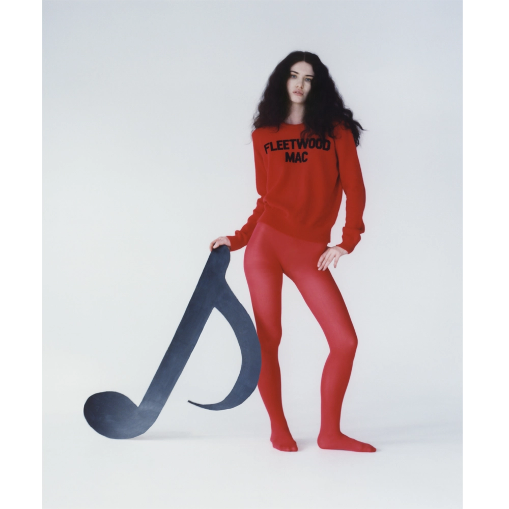 A model wearing the red Fleetwood Mac knit jumper, shown against a plain light background whilst holding a handmade black music note.