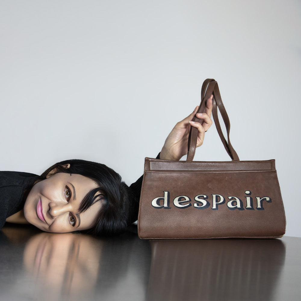 Artist Gillian Wearing lies on a reflective dark table, holding up a brown leather handbag with the word 'despair' spelled out in large printed letters on the front, against a plain light grey background.
