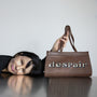 Artist Gillian Wearing lies on a reflective dark table, holding up a brown leather handbag with the word 'despair' spelled out in large printed letters on the front, against a plain light grey background.