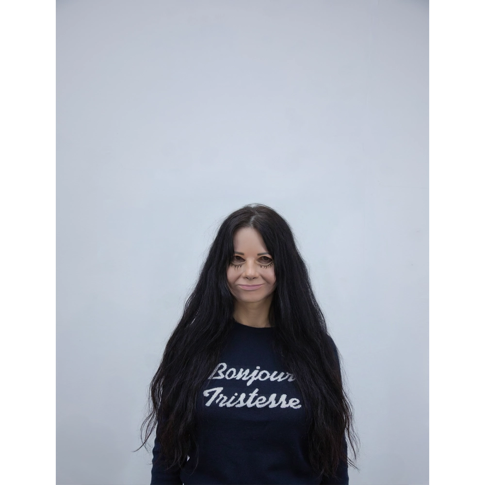 Artist Gillian Wearing, wearing a dark navy sweatshirt with 'Bonjour Tristresse' printed in white script across the chest, set against a plain light grey background.