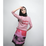 Artist Gillian Wearing wearing a light pink long-sleeve sweatshirt with 'Bonjour Tristesse' written in red script across the chest, paired with a pink, red, and black patterned skirt, posing against a plain light grey background.