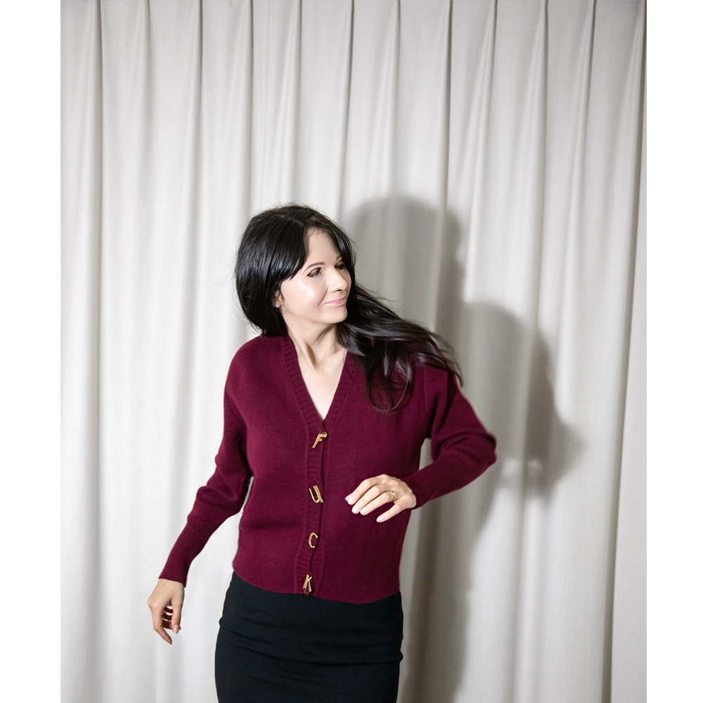 Artist Gillian Wearing wearing a burgundy knit cardigan with gold buttons and a black skirt, standing in front of a light beige curtain backdrop wearing a mask.