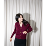 Artist Gillian Wearing wearing a burgundy knit cardigan with gold buttons and a black skirt, standing in front of a light beige curtain backdrop wearing a mask.