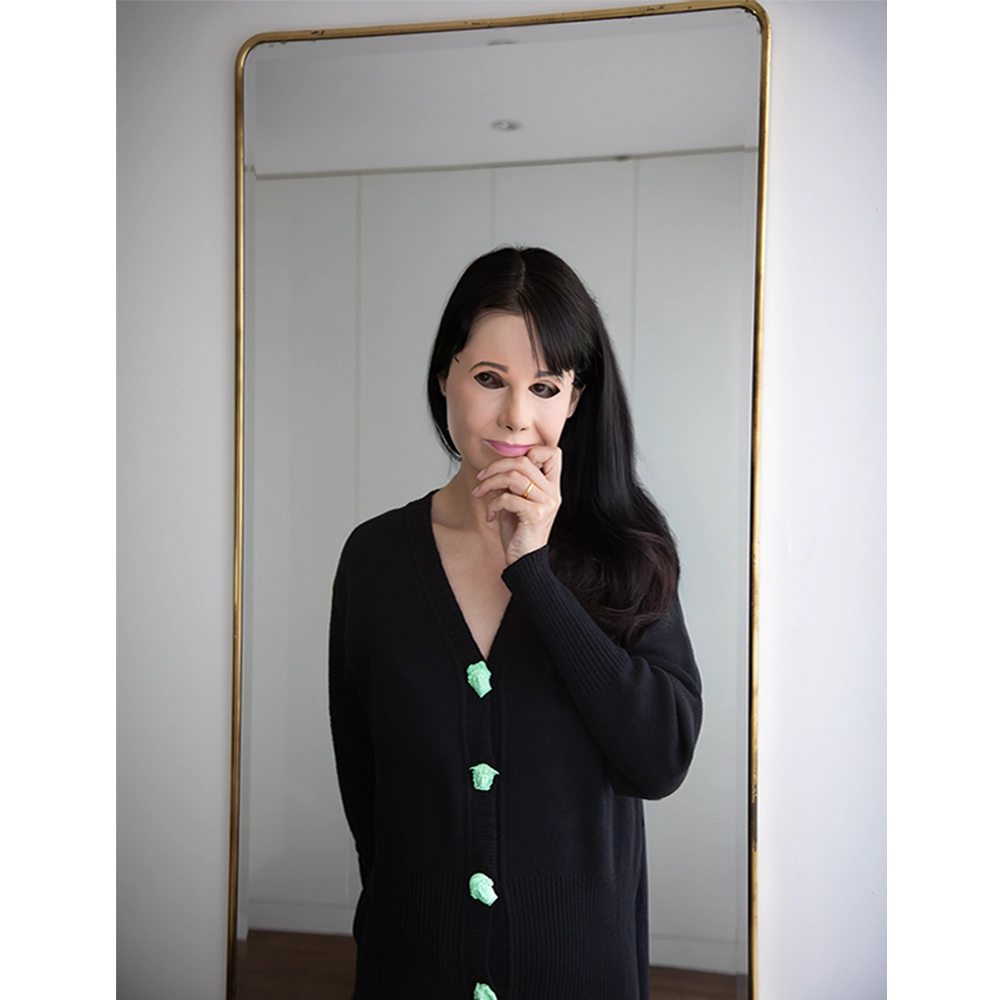 Artist Gillian Wearing wearing the Greek Mythology Cardigan, in front of a mirror wearing a mask to cover her face. 