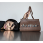 Artist Gillian Wearing lies on a reflective dark table, holding up a brown leather handbag with the word 'despair' spelled out in large printed letters on the front, against a plain light grey background.
