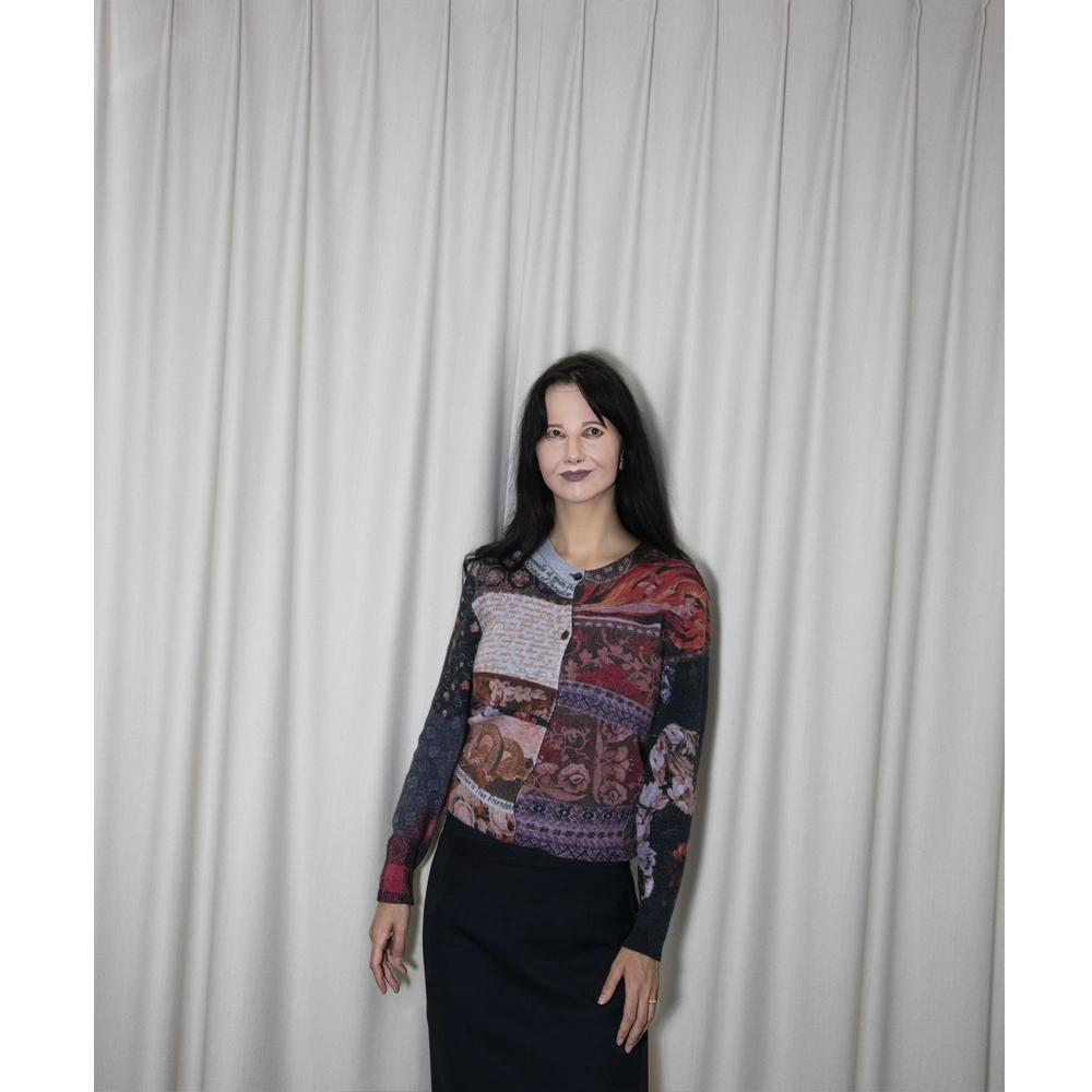 Artist Gillian Wearing stands in front of a white curtain backdrop wearing a long-sleeve William Blake patchwork cardigan , paired with a dark skirt.