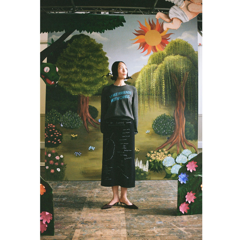 Model wearing the "Make Friends With Chaos" knit and Notes On Radical Living skirt against a faux garden of eden backdrop.