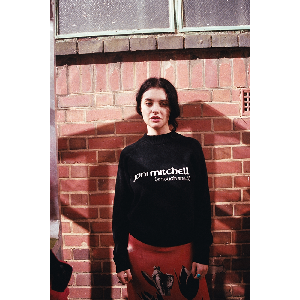 Model wearing a navy sweater with 'joni mitchell (enough said)' text against a brick wall.