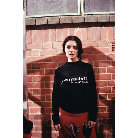 Model wearing a navy sweater with 'joni mitchell (enough said)' text against a brick wall.