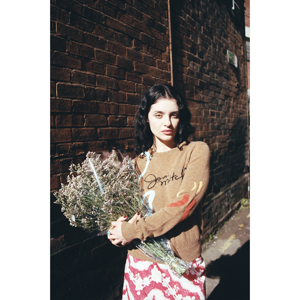 Woman wearing brown 'Joni Mitchell' jumper holding flowers against a brick wall