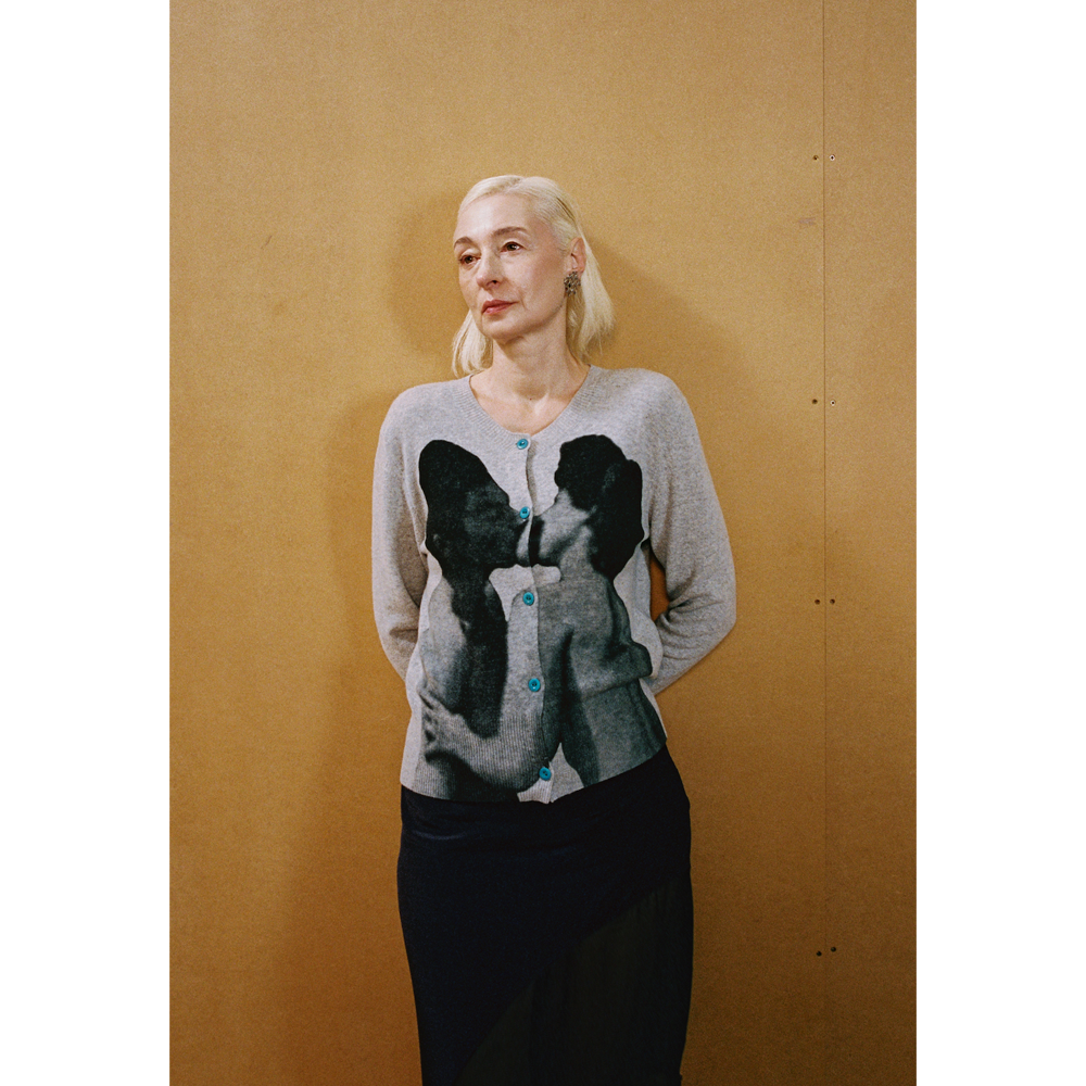 Close up of a lady wearing the grey Kissing Cardigan and navy Delusion Skirt leaning against a wooden wall
