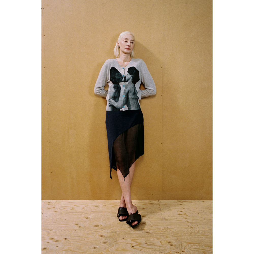 Lady wearing the grey Kissing Cardigan and navy Delusion Skirt leaning against a wooden wall