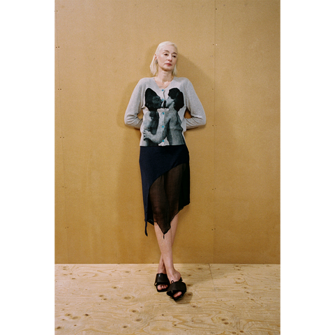 Lady wearing the grey Kissing Cardigan and navy Delusion Skirt leaning against a wooden wall