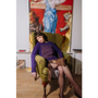 Woman in a purple Manifesto jumper by HADES Wool sitting on a green armchair with a large painting in the background