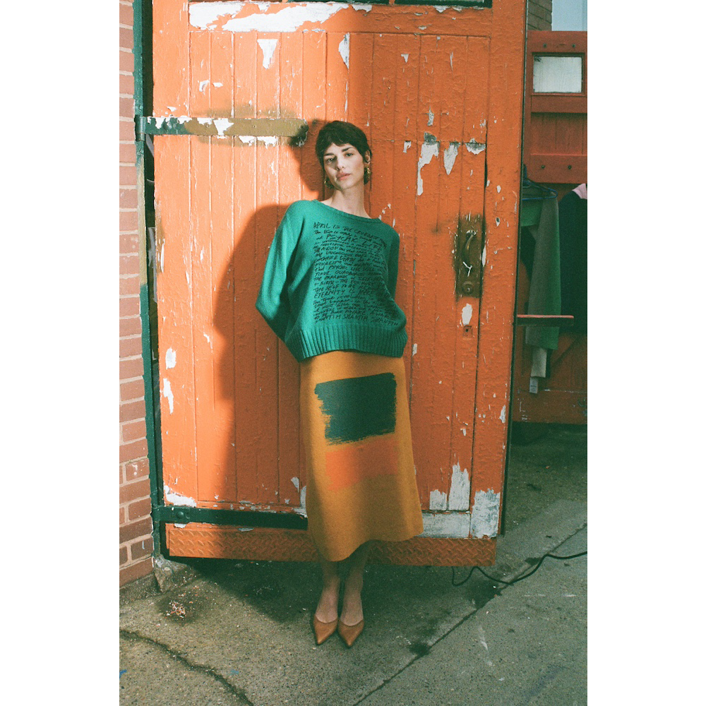 Woman in green HADES manifesto jumper and yellow Second Half of Life skirt standing against a orange door.