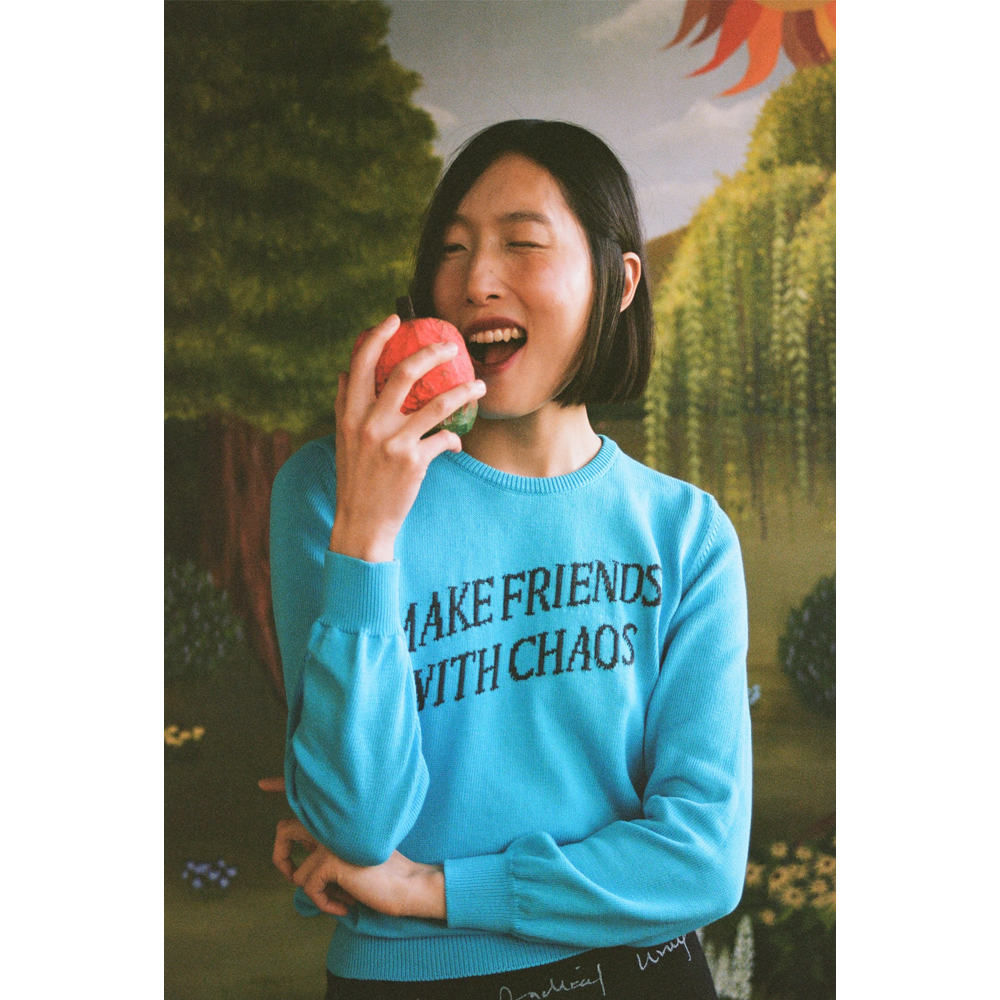 Model wearing a "Make Friends With Chaos" knit holding a faux apple in a faux garden of eden.