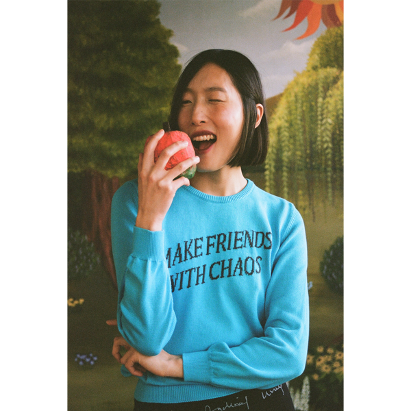 Model wearing a "Make Friends With Chaos" knit holding a faux apple in a faux garden of eden.