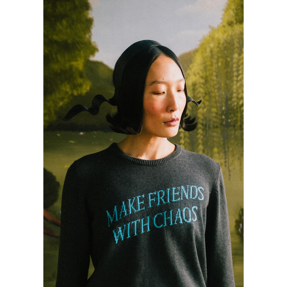 Model wearing a "Make Friends With Chaos" knit against a faux garden of eden.